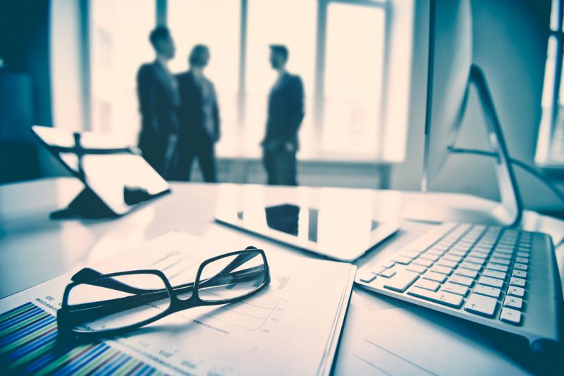 Modern devices, documents, eyeglasses on the foreground, unrecognized businessmen on the background